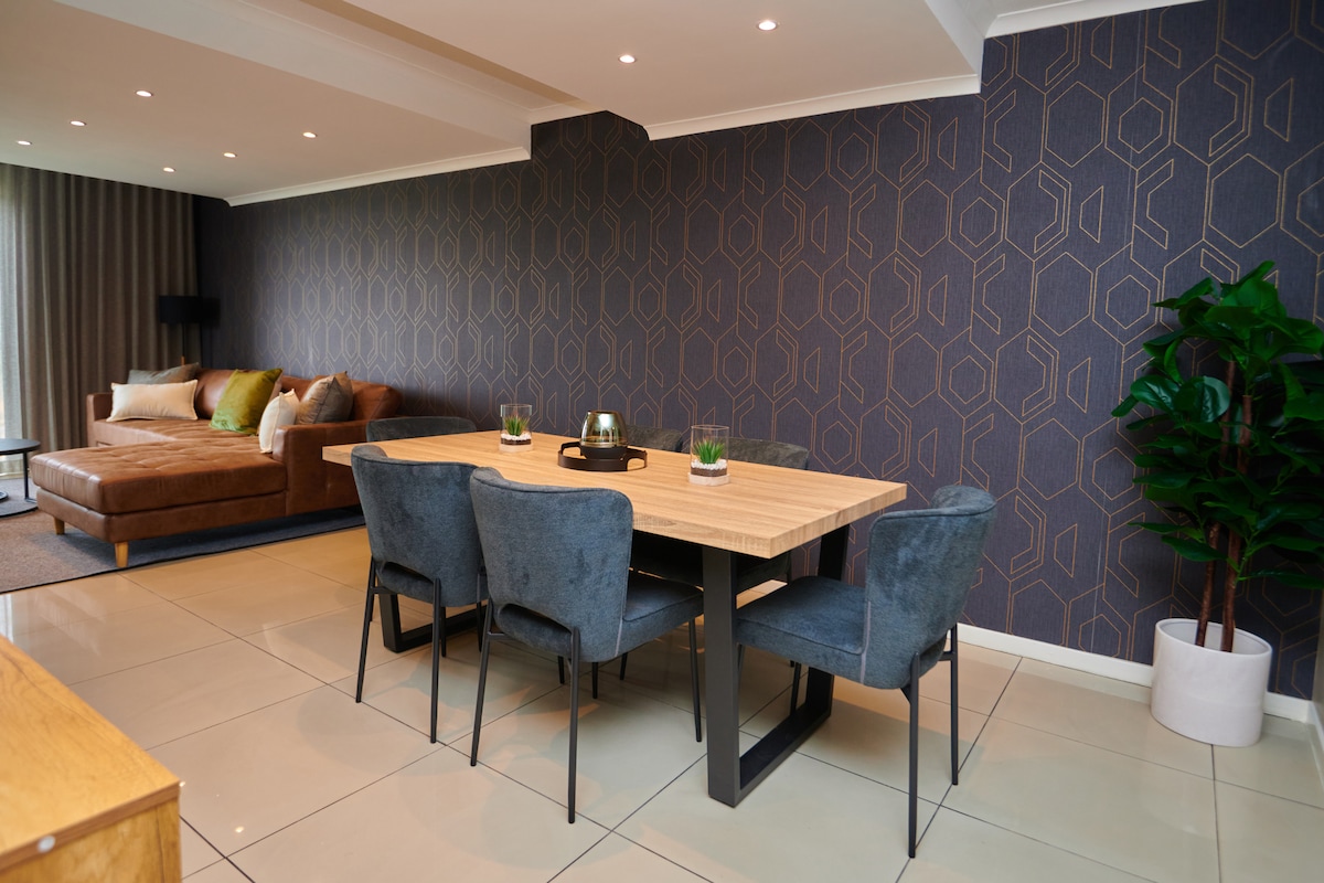 A modern dining area features a rectangular wooden table surrounded by four charcoal-grey upholstered chairs. The backdrop consists of a geometric patterned wallpaper in deep blue and gold. A plush sectional sofa in a warm brown tone is positioned nearby, creating a cozy atmosphere.