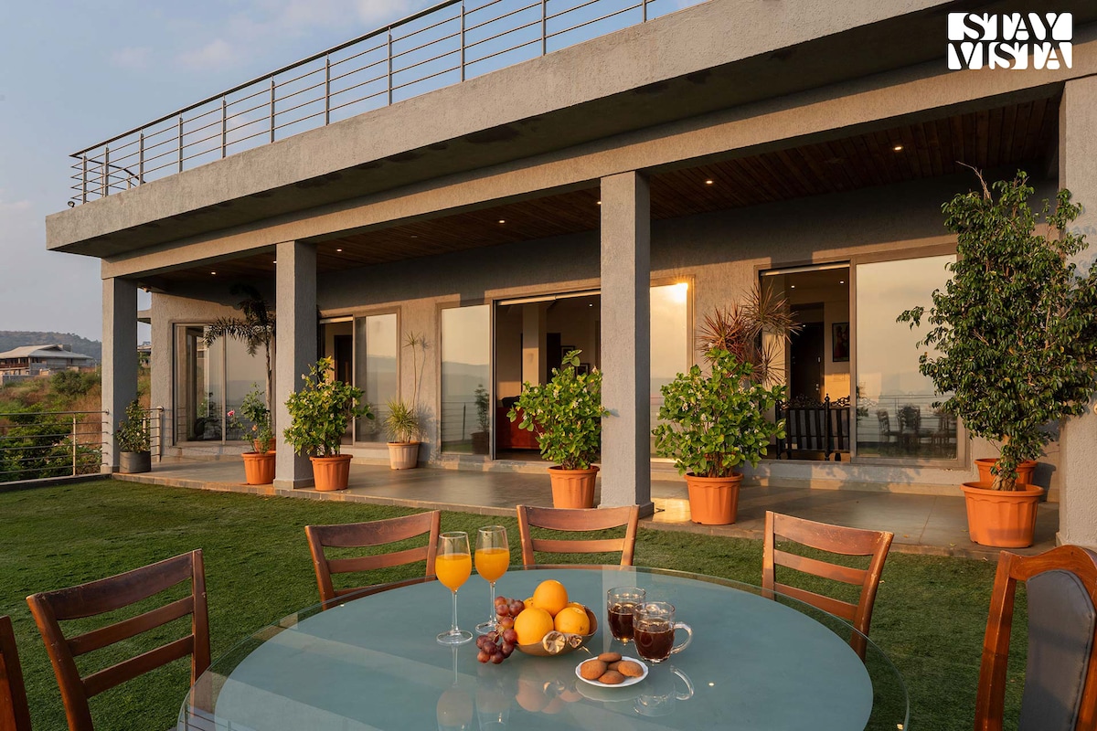 An outdoor dining area features a circular table set with refreshing beverages and a selection of fruits. Potted plants enhance the space, while large sliding doors lead to bright indoor areas, creating a seamless connection with nature and the scenic surroundings.