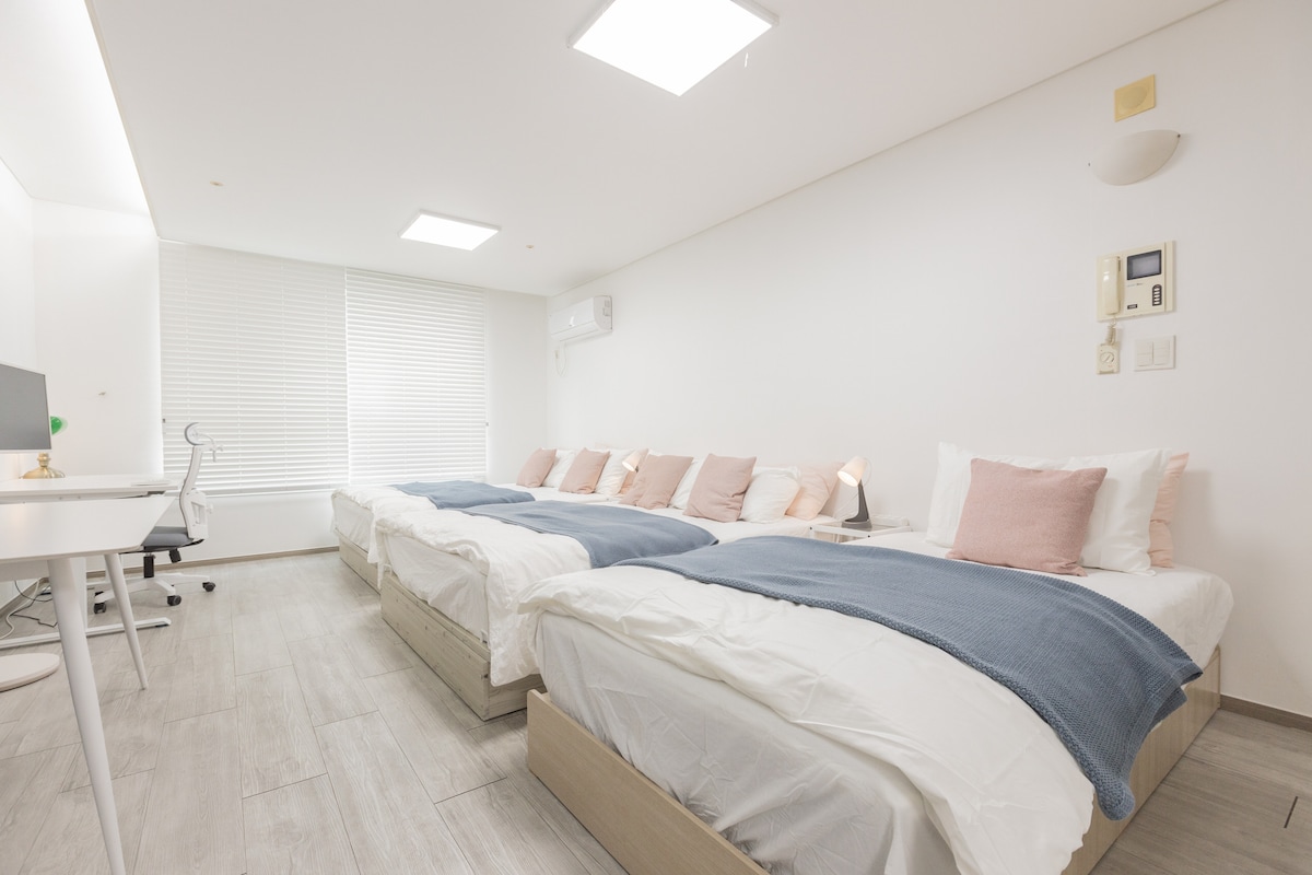 The sleeping area features two spacious queen beds and a single bed, elegantly arranged with soft linens and decorative pillows. A desk with a chair is positioned nearby, complemented by natural light from large windows covered with light blinds.