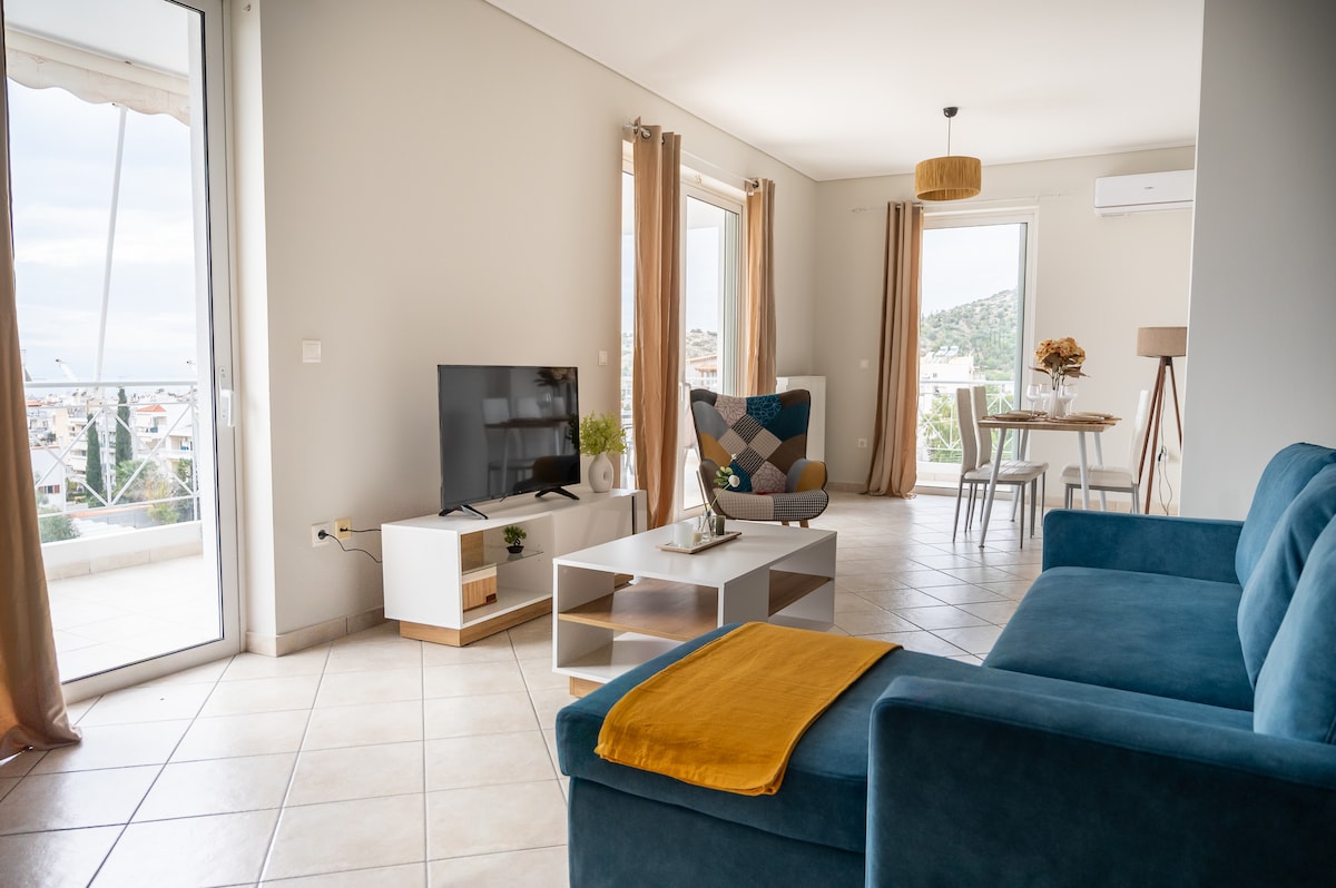 A spacious living area is presented with a comfortable blue sofa and a light wood entertainment unit. A flat-screen TV is positioned on the unit. Natural light fills the space through large glass doors leading to a balcony, enhancing the airy feel.