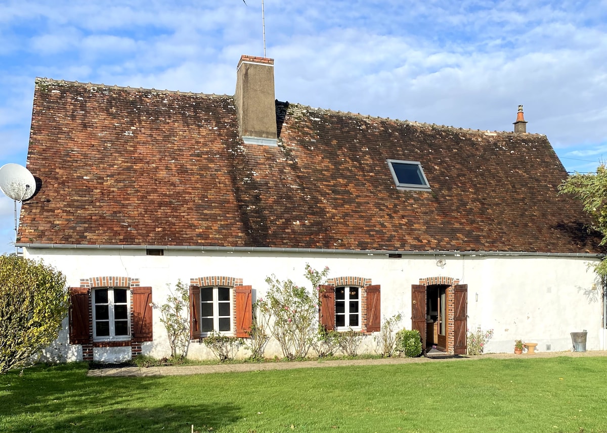 A charming farmhouse is depicted, featuring a sloping roof and a light-colored exterior. Several windows are framed with wooden shutters, while a spacious lawn with greenery surrounds the house. A small patio area is visible at the entrance, enhancing the outdoor space.