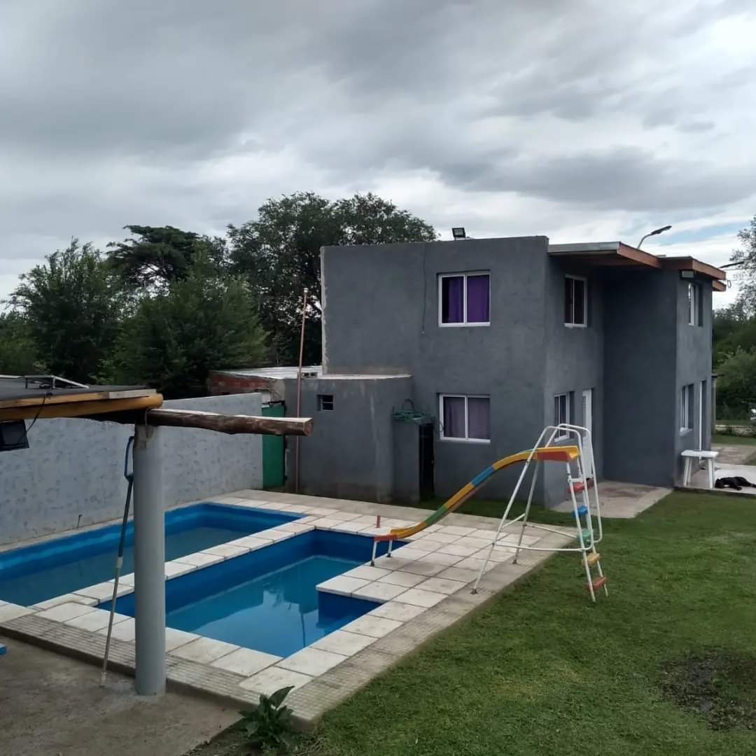 A two-story house is shown adjacent to a swimming pool, which features a small slide. The structure is painted in a muted grey tone, with multiple windows displaying purple accents. Surrounding the house, green grass and trees provide a natural setting.