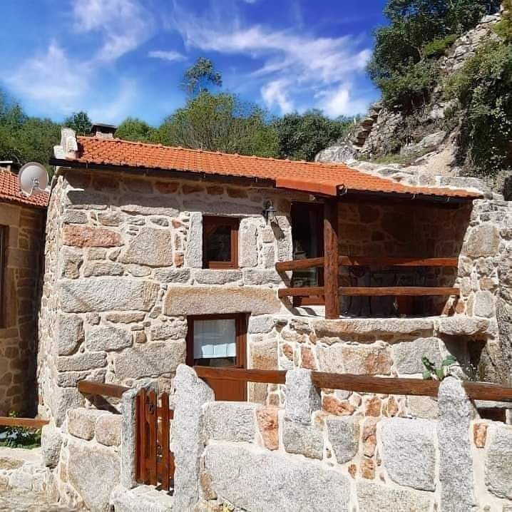 A stone cottage stands nestled against a natural backdrop, featuring rustic elements and a distinctive red-tiled roof. Wooden railings are visible on the porch, while the surrounding landscape shows greenery under a clear blue sky.