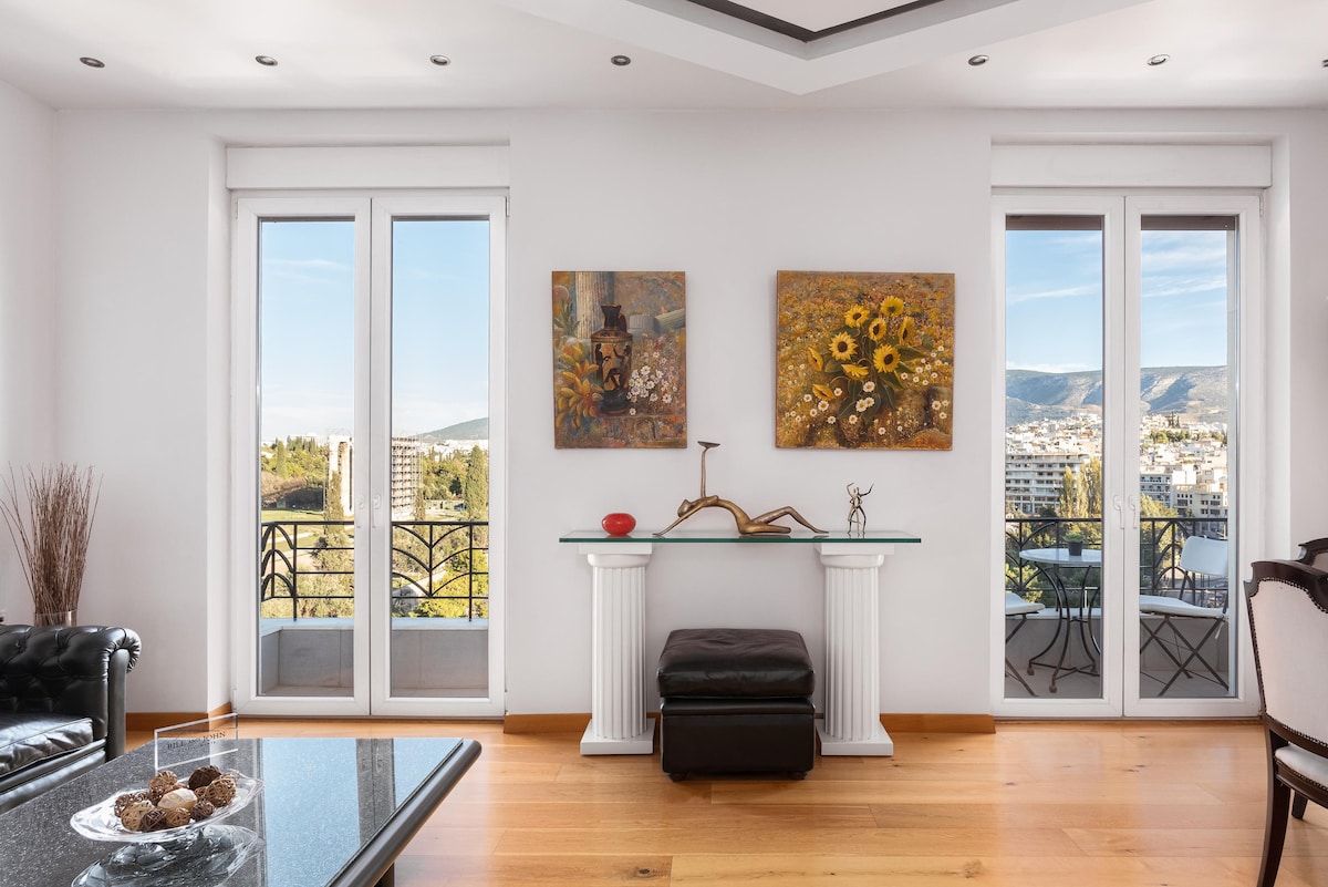 A bright living area features large glass doors providing access to a balcony with scenic views. Two framed artworks adorn the white walls, complemented by a stylish console table and a modern seating area, creating an open and inviting ambiance.
