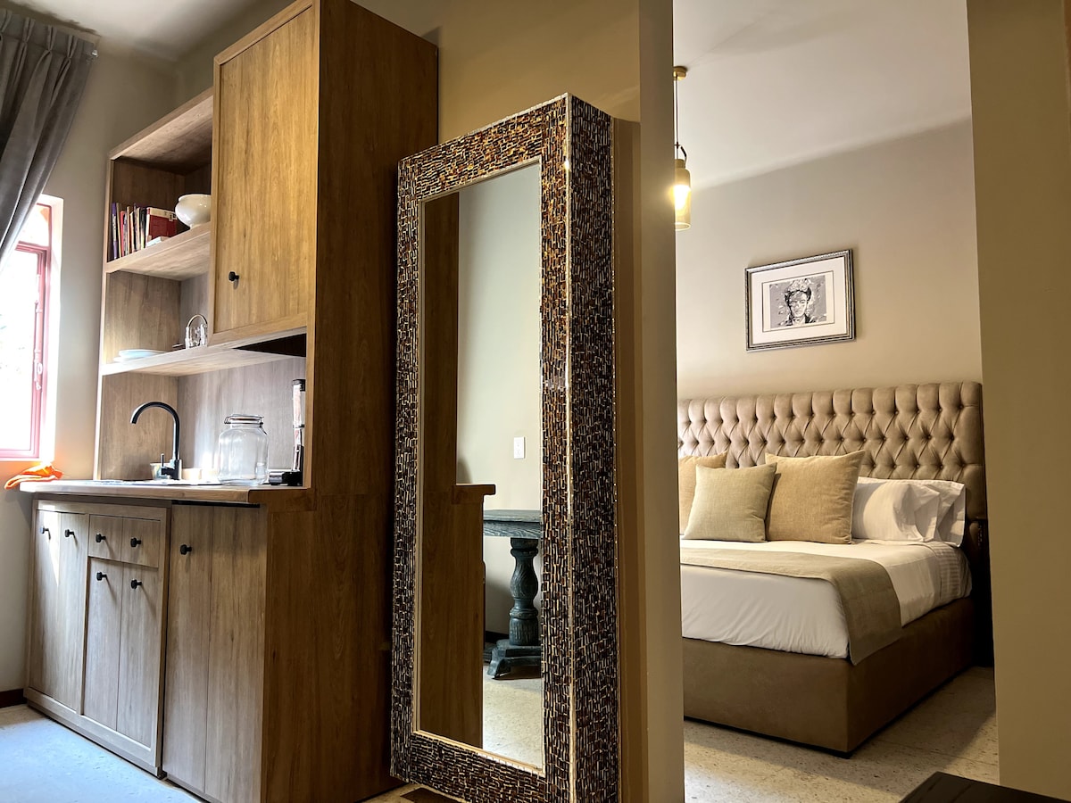 A well-appointed studio features a spacious layout with a kitchen area on one side. A large, decorative mirror reflects the cozy sleeping area, which includes a comfortable bed with a tufted headboard. Natural light enters through a window, complementing the warm wood tones.