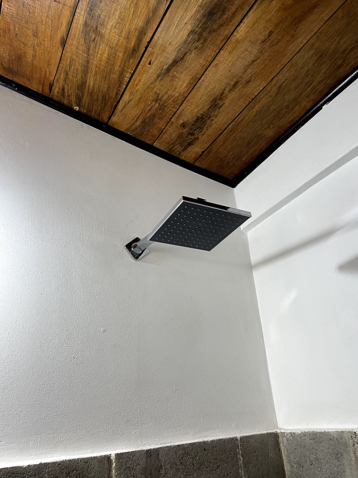 A modern shower head with a square design is mounted on a clean white wall. The wall is complemented by a warm wood-paneled ceiling, creating a contrast with the sleek metal fixture.