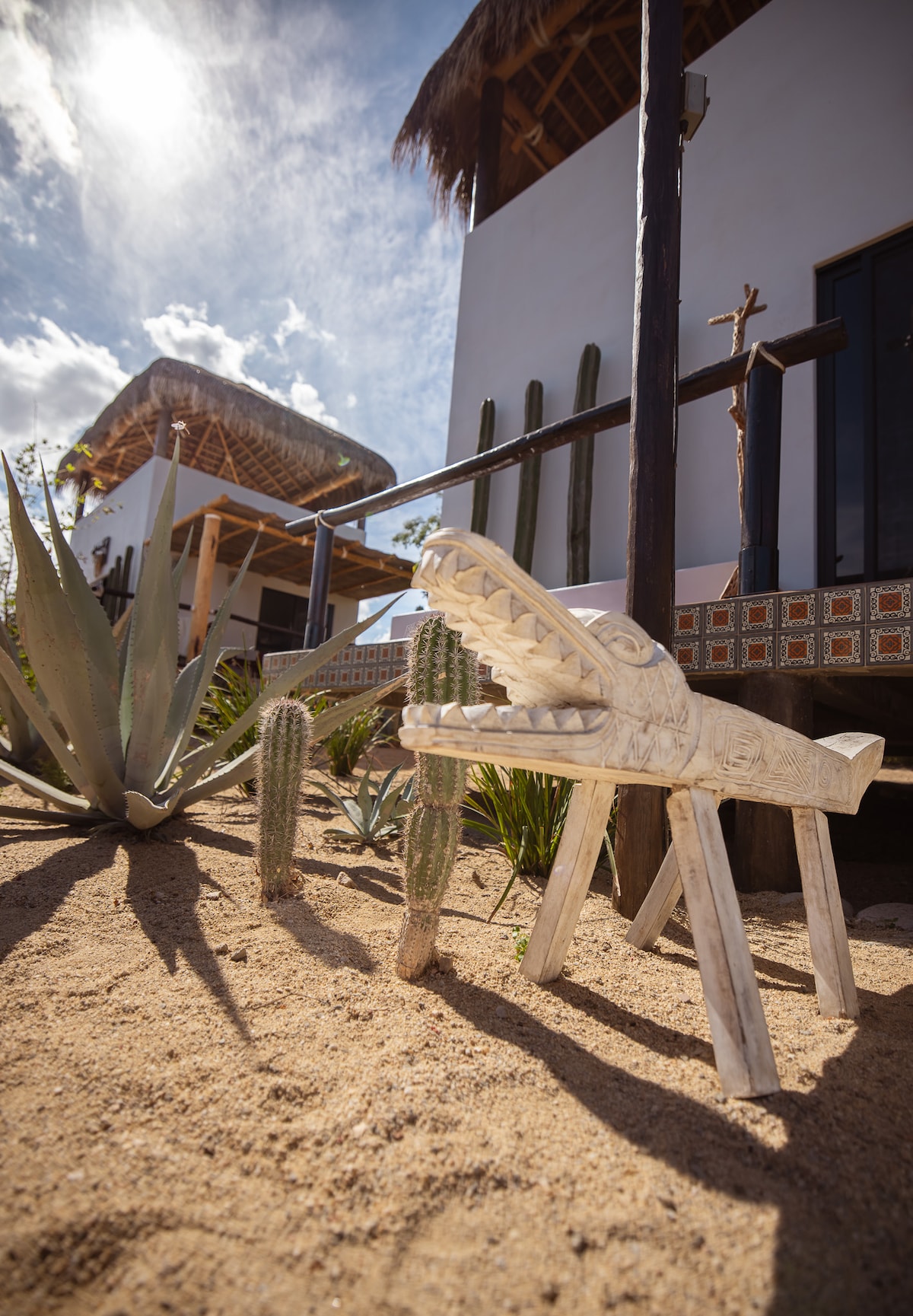A carved stone animal sculpture is positioned in a sandy area, surrounded by cacti and desert plants. In the background, the rooftops of the nearby houses are visible under a bright sky, adding to the natural ambiance of the outdoor space.
