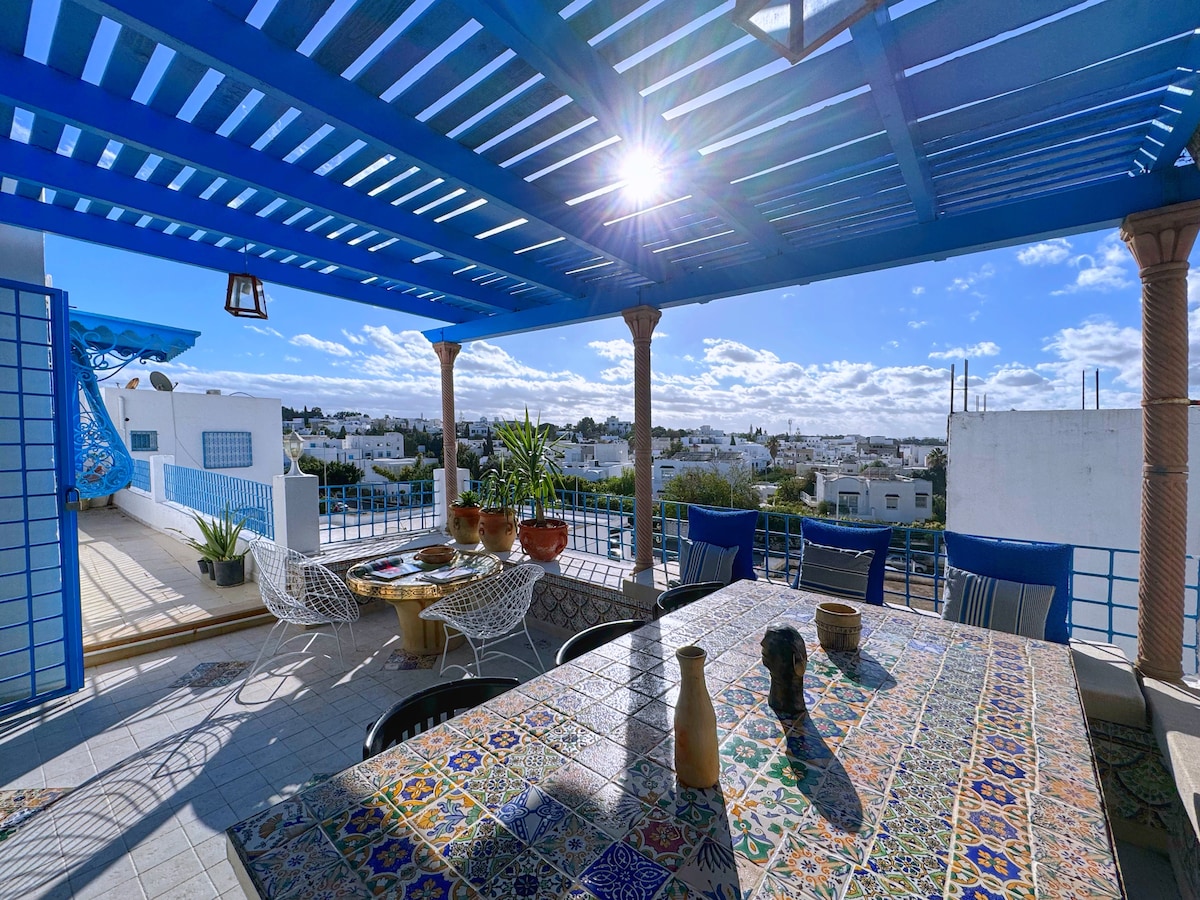 A spacious terrace is shown, featuring a decorative tiled table surrounded by white woven chairs. Potted plants add a touch of greenery. The blue pergola overhead filters sunlight, casting shadows on the floor. A panoramic view of Sidi Bou Said's rooftops is visible in the background.
