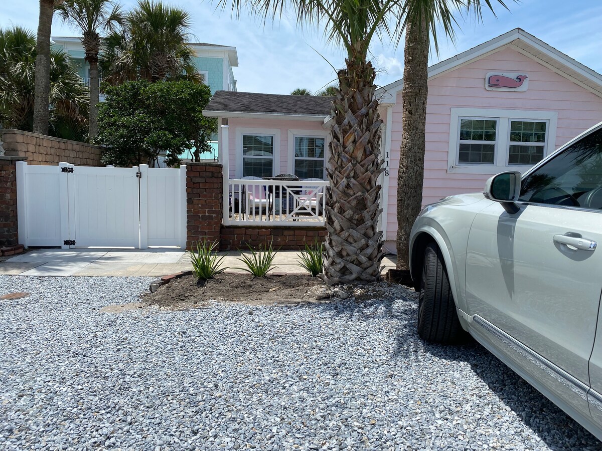 The exterior of the cottage is featured, showcasing a charming pink facade framed by lush greenery. A white gate leads to a well-manicured area, with gravel pathways and tropical plants visible. A parked vehicle is positioned on the right side of the image.