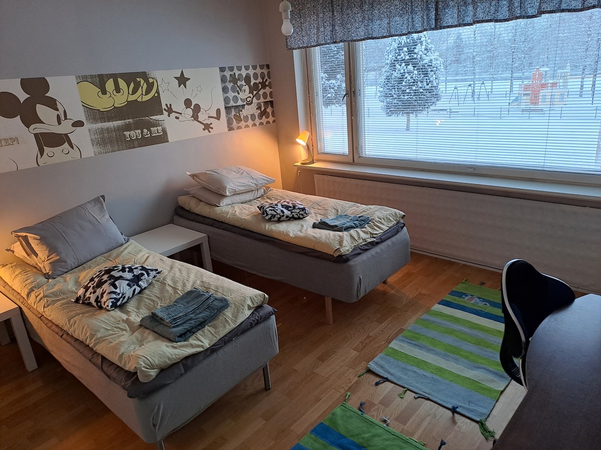 A bedroom features two spacious beds topped with soft linens and throws. A colorful rug lies on the wooden floor, while a window reveals a snowy outdoor scene with playground equipment in the distance. A small desk sits nearby, offering a functional workspace.