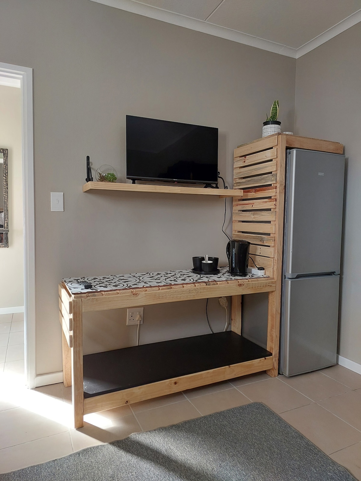 A wooden console table is positioned against a neutral wall, featuring a flat-screen TV mounted above a shelf. A modern fridge stands adjacent, with a kettle and other items placed on the table, offering convenience for guests.
