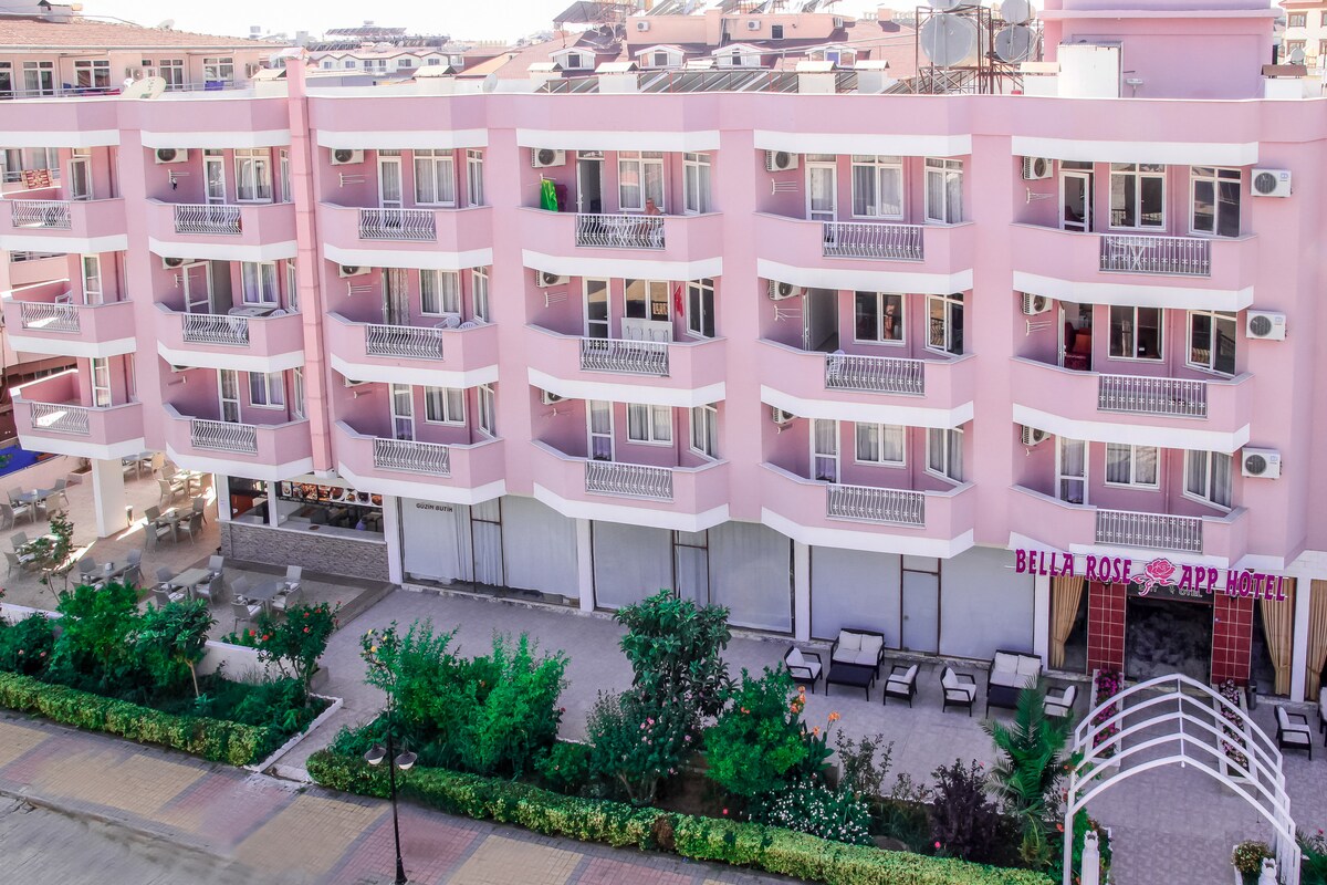 A pink, three-story building features multiple balconies with sliding glass doors. The ground level showcases an outdoor seating area surrounded by landscaped greenery. An arched entrance leads to the property, highlighting the name 'BELLA ROSE APP HOTEL' prominently at the base.