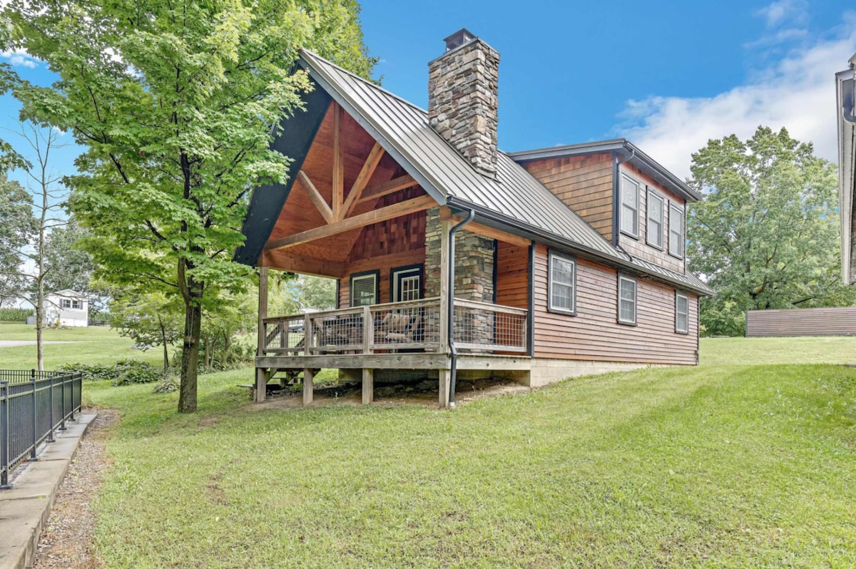 A cabin exterior is presented under a clear blue sky, showcasing a covered porch supported by a stone and wooden framework. Lush green grass surrounds the structure, with large trees providing shade in the foreground and background.