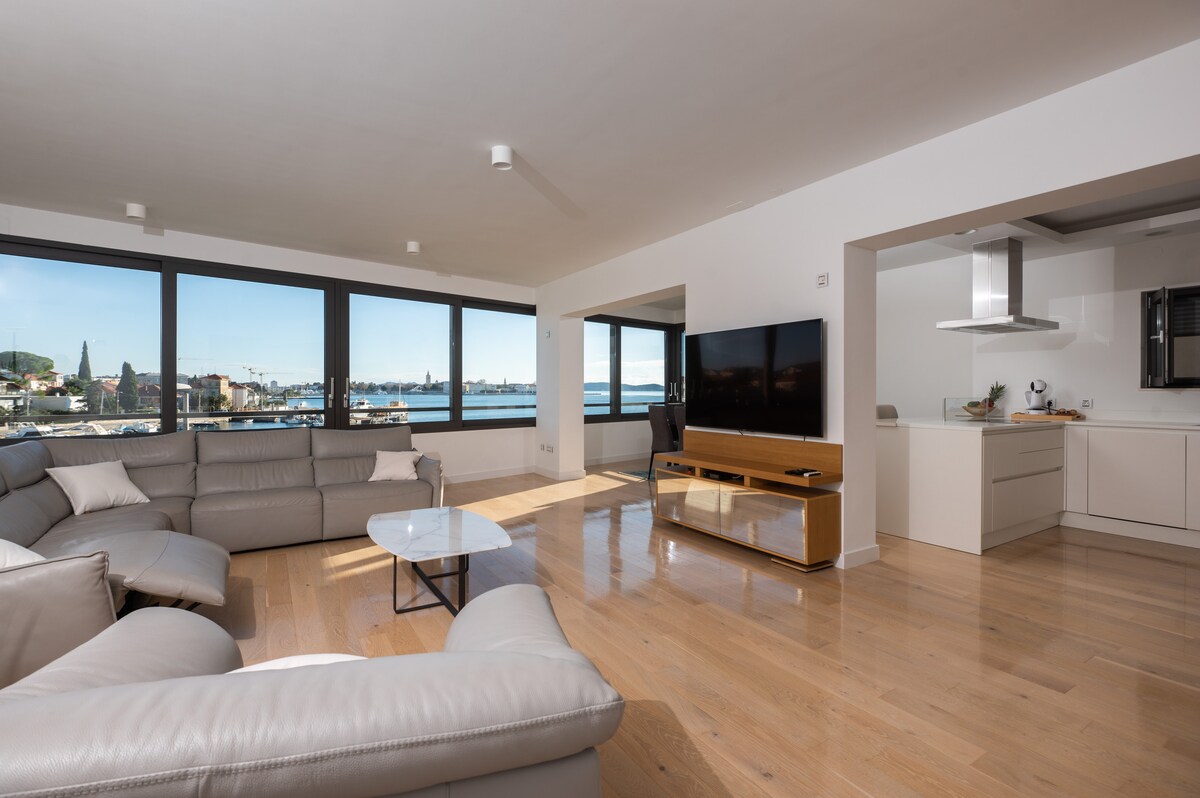 A modern living area features a large sectional sofa arranged around a low coffee table. Floor-to-ceiling windows allow for abundant natural light and views of the waterfront. An open kitchen with sleek cabinetry is visible in the background, enhancing the space's contemporary design.