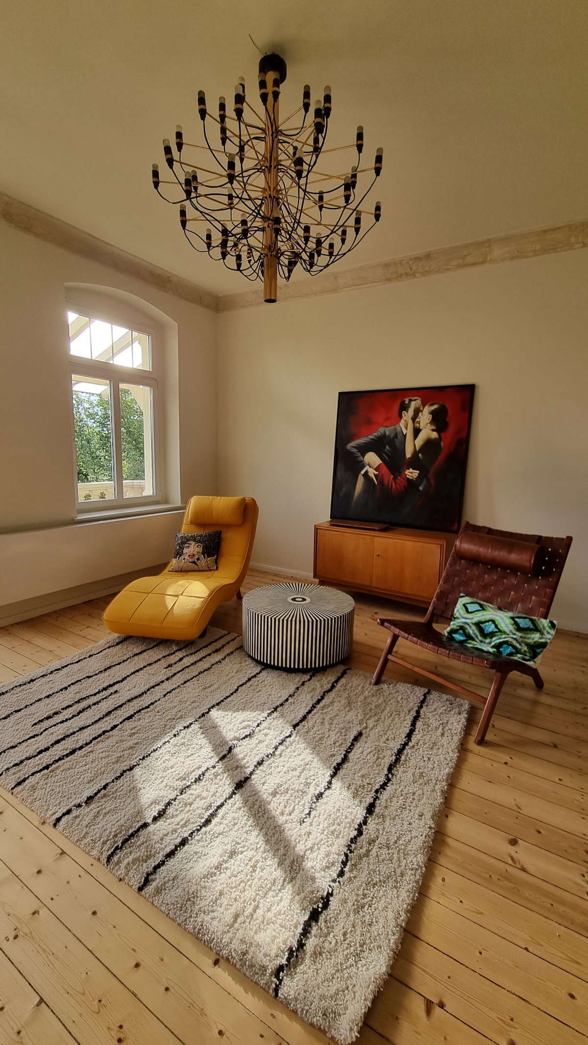 A well-lit living area features a cozy lounge chair and a wooden rocking chair, positioned on a soft area rug. A round coffee table complements the seating, while a vibrant wall painting adds visual interest. The space is enhanced by a decorative chandelier and natural light.