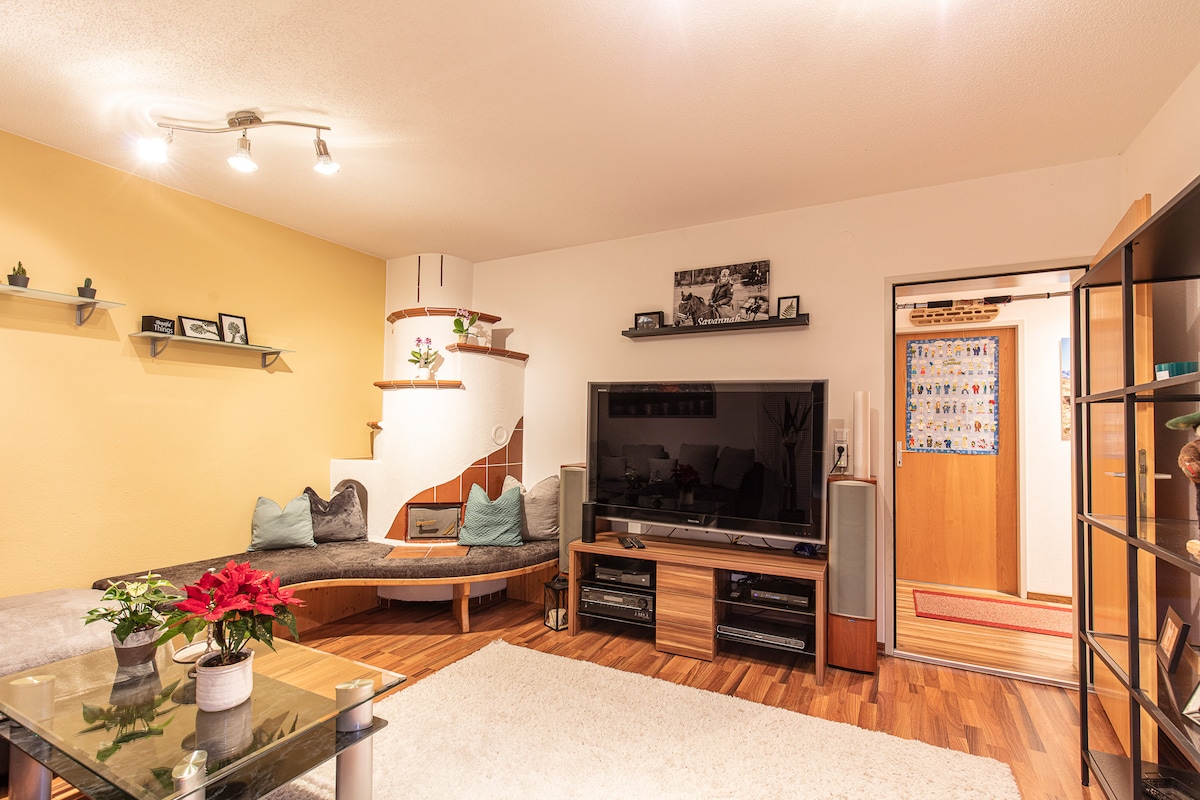 The living area features a comfortable curved seating arrangement with decorative cushions and a glass coffee table at the center. A large television is positioned against a light wall, while shelves display various plants and photographs. Warm lighting creates a welcoming ambiance.