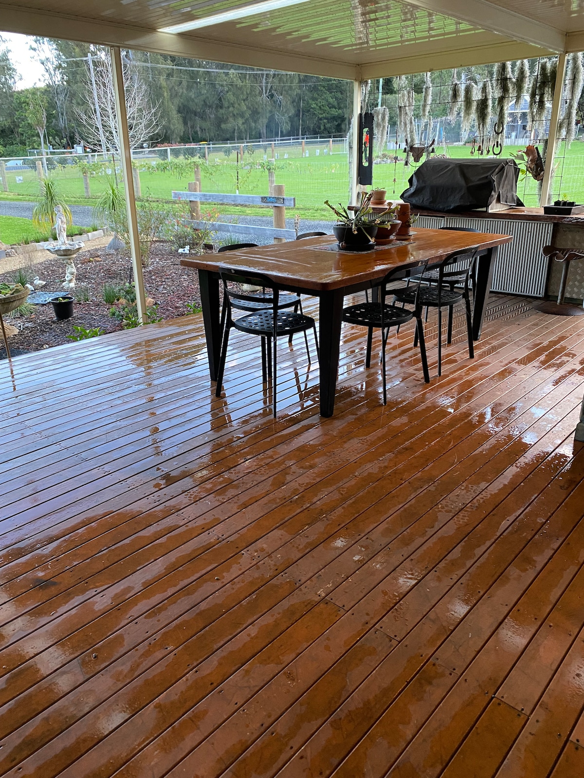A covered outdoor deck features a polished wooden floor, with a dining table made of dark wood and four matching chairs. A barbecue grill is positioned nearby, and the lush green landscape is visible just beyond the deck, enhancing the outdoor experience.