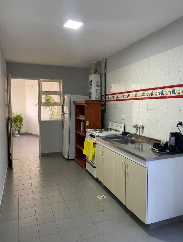 A functional kitchen is showcased, featuring a stainless steel sink and a stove. Cabinets line the walls, providing storage solutions. A refrigerator is visible, and a bright yellow towel adds a touch of color. Natural light filters in through a window at the far end.