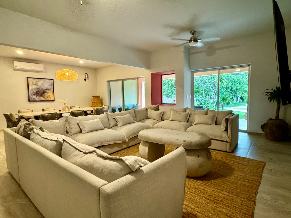 A spacious living area is furnished with a large, light-colored sectional sofa arranged around a round coffee table. A textured area rug is placed underneath, while large sliding glass doors lead to a green outdoor space, providing ample natural light.