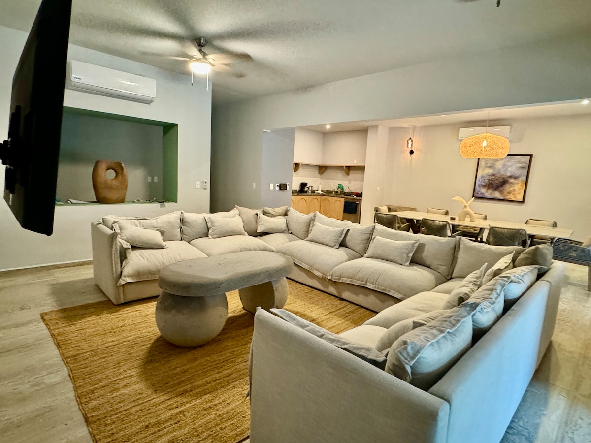 A spacious living area features a large, circular, cushioned sofa arranged around a stone coffee table. Natural light enters from nearby windows, enhancing the open layout. The kitchen and dining area are visible in the background, promoting a communal atmosphere.