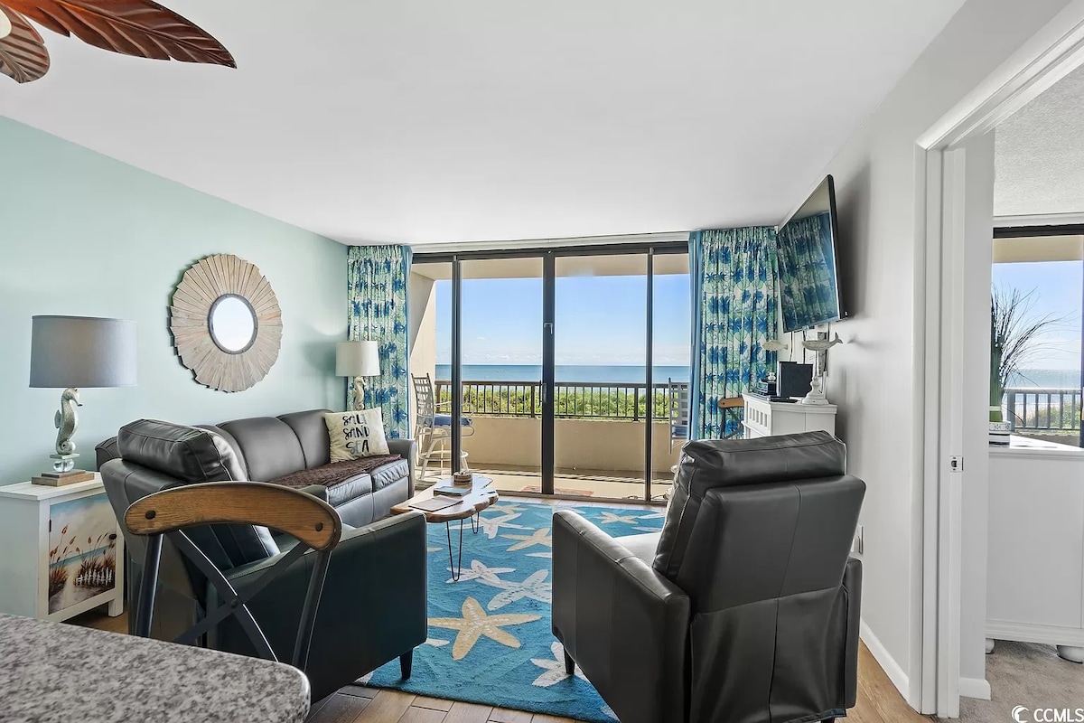 A comfortable living area is showcased with a leather sofa and two reclining chairs. Floor-to-ceiling windows provide ocean views, while vibrant blue décor complements the space. A patterned area rug lies on the floor, and a TV is mounted on the wall opposite the seating.