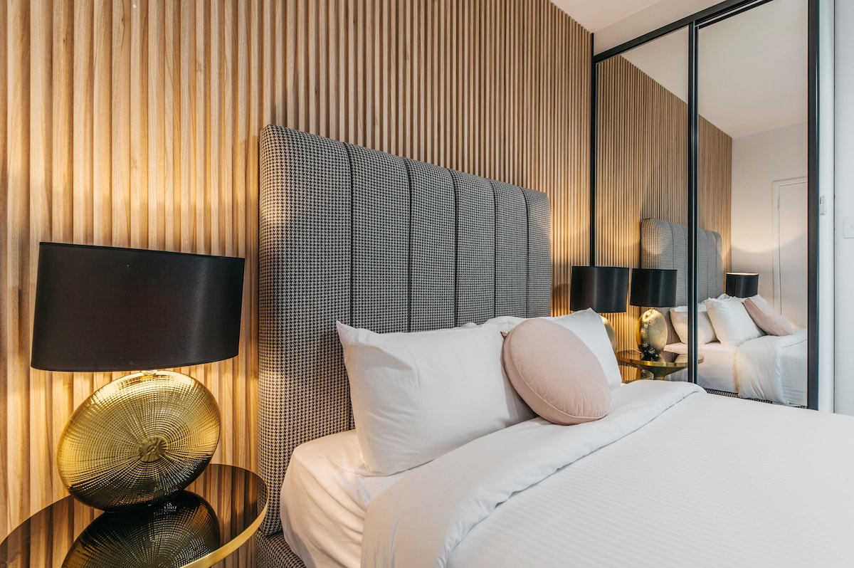 The bedroom features a modern headboard with a textured gray design. A plush bed is dressed in white linens and a round decorative pillow. Warm light is emitted from stylish lamps on each side, and a mirrored wardrobe reflects the calming ambiance.