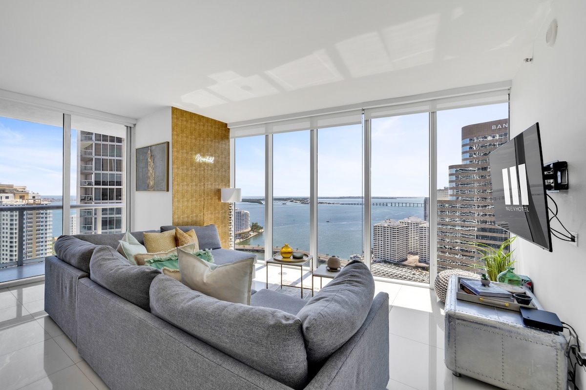The living room showcases a light-filled space with floor-to-ceiling windows, providing expansive views of Biscayne Bay. A large, comfortable sectional sofa is positioned nearby, complemented by a TV mounted on the wall. The sleek décor emphasizes a modern aesthetic, enhancing the inviting atmosphere.
