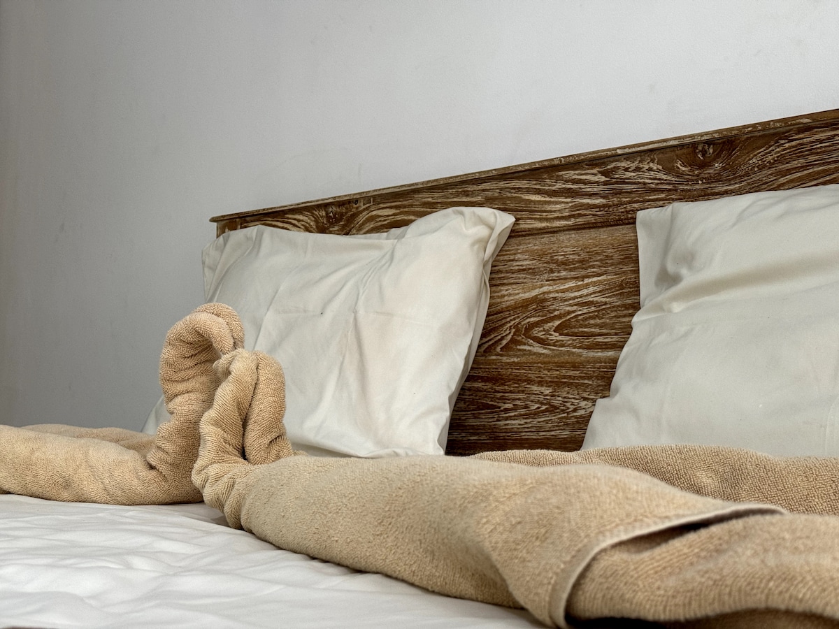 A spacious bed is adorned with two white pillows and a soft beige blanket casually draped over the edge. The natural wood headboard adds a warm touch to the minimalist sleeping area, complemented by a light neutral wall.