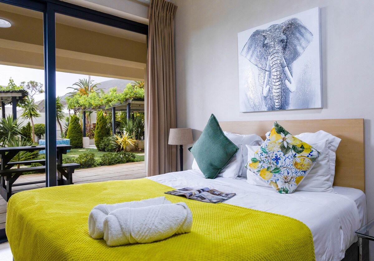 A well-lit sleeping area features a spacious bed adorned with a bright yellow bedspread and decorative pillows. A framed elephant artwork hangs above the bed. Sliding glass doors open to a garden view with seating areas and lush greenery visible outside.