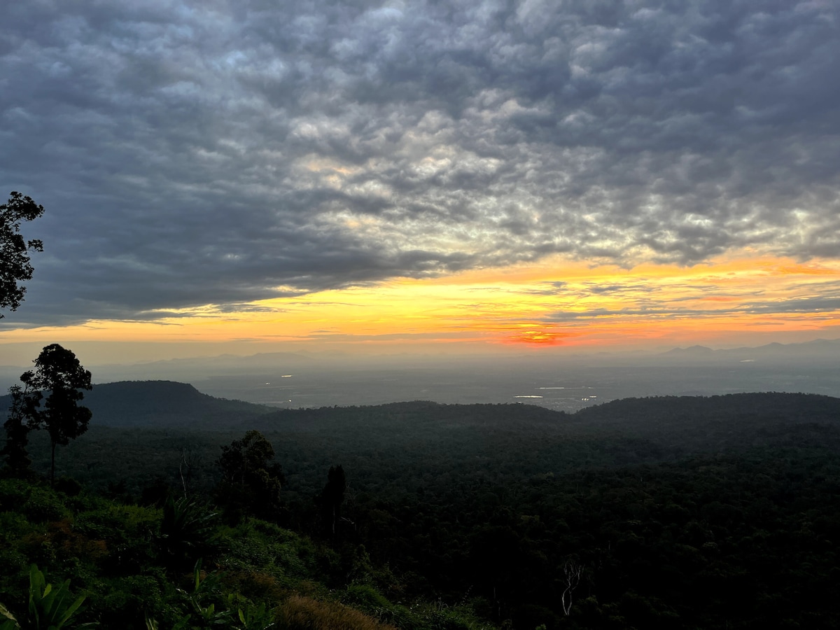 A wide panoramic view of a vast mountain landscape is depicted, showcasing a sunset that fills the sky with warm hues of orange and yellow against a backdrop of clouds. Lush greenery covers the rolling hills, providing a sense of serene nature.