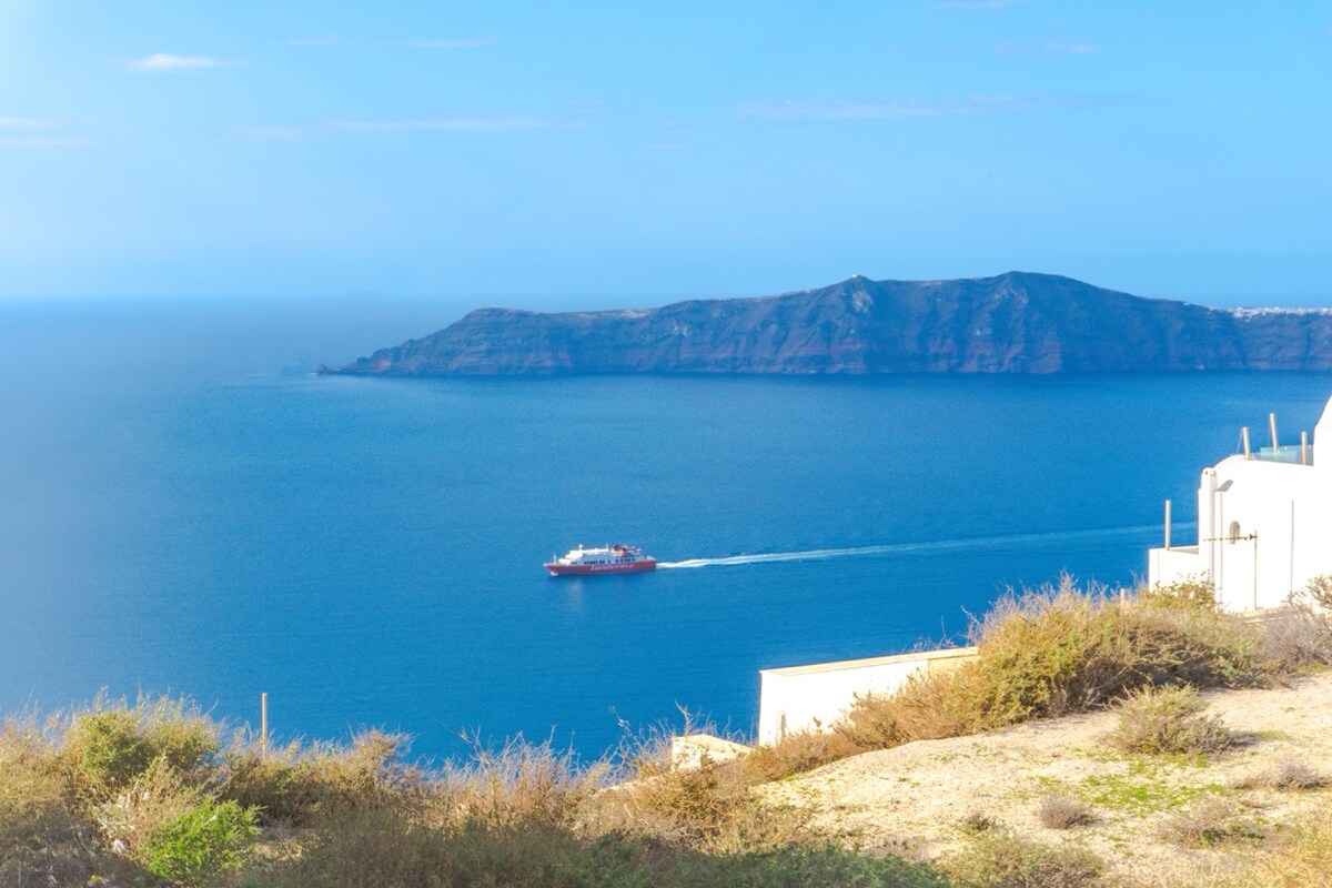 A view overlooking a calm sea, with a red ship gliding across the water towards a distant island. The landscape is characterized by gentle hills and sparse vegetation, showcasing the natural beauty of the coastline.