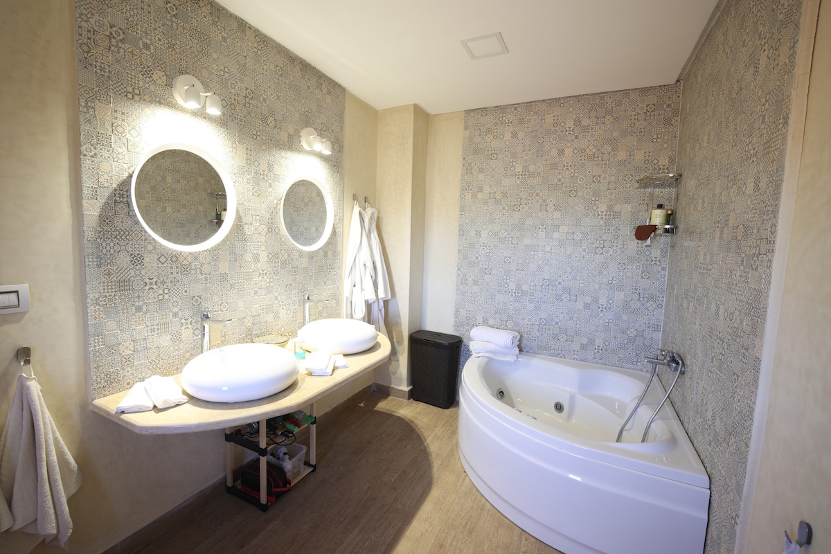 A well-appointed bathroom features two modern sinks positioned on a sleek countertop. Above each sink, round mirrors reflect soft lighting from wall-mounted fixtures. A curved bathtub is situated in the corner, complemented by decorative tiles that enhance the overall design.