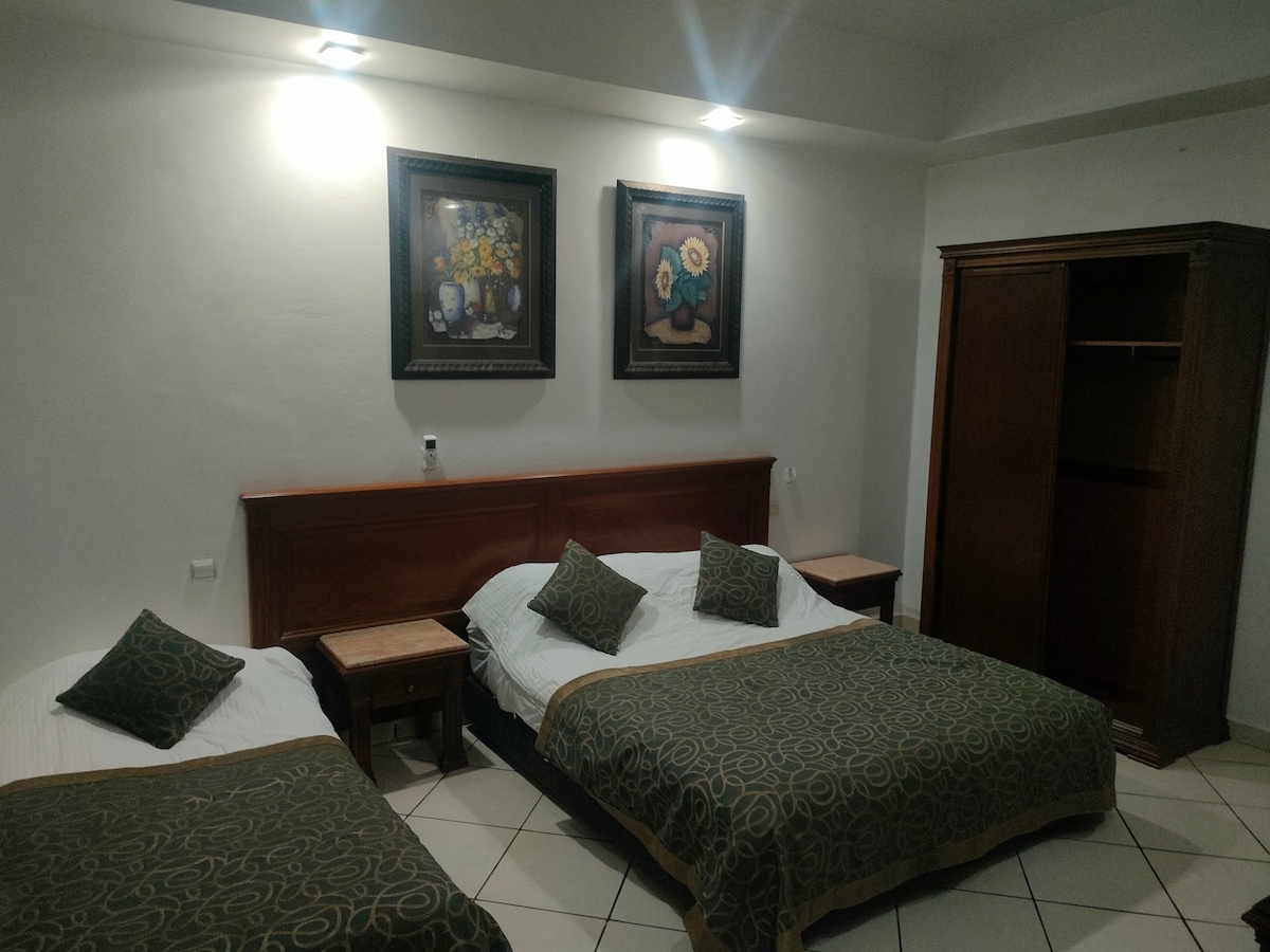 A well-furnished room is showcased with two spacious beds, each adorned with patterned bedspreads. Decorative artwork featuring flowers hangs above the beds. A wooden wardrobe stands nearby, complemented by two bedside tables that provide additional convenience. Wall lighting enhances the room's overall brightness.