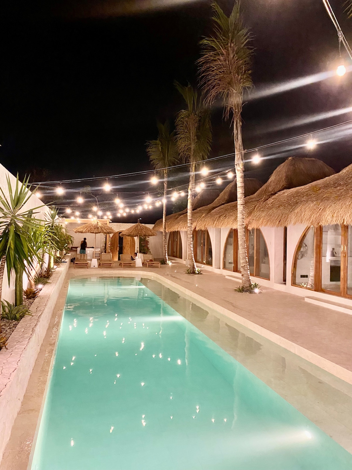 The pool area is bathed in soft lighting, with a refreshing turquoise pool reflecting the serene ambiance. Surrounding palm trees and thatched-roof structures create a tropical vibe, while string lights add a cozy glow, enhancing the inviting atmosphere for evening relaxation.