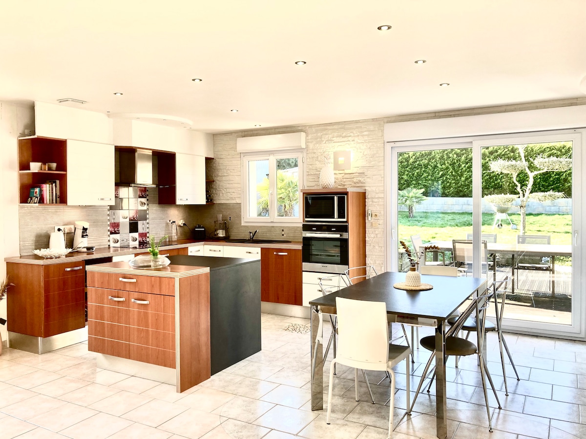 A modern kitchen is equipped with wooden cabinetry and stainless steel appliances. A central island with seating is present, alongside a dining table surrounded by chairs. Natural light floods the space through large sliding doors that lead to the garden.