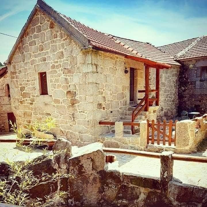 A charming stone house with a rustic facade is depicted. A wooden porch and steps lead to the entrance, while a small fence surrounds the front yard. The surrounding area features cobbled pathways and traditional architecture, enhancing the village ambiance.
