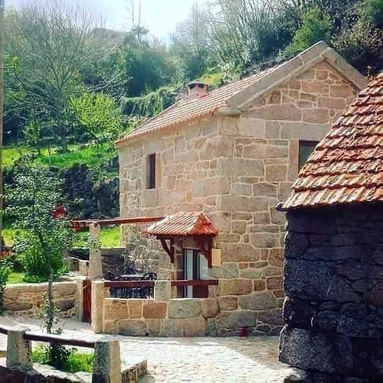 A stone house with a red-tiled roof is nestled in a lush green landscape. The structure features large windows and a welcoming porch adorned with patio furniture. Surrounding trees and hillside create a tranquil outdoor environment.