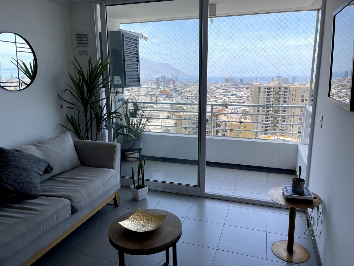 A modern living area features a light-colored couch paired with a small round table. Floor-to-ceiling sliding glass doors provide access to a balcony with a city view, while plants are placed thoughtfully in the space. A circular mirror adds depth to the room.