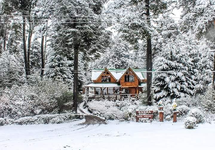 A two-story wooden cabin is surrounded by a snowy landscape, with snow-covered trees creating a serene setting. The cabin features a sloped roof and a welcoming porch. A sign in front identifies the property, partially obscured by the snow.