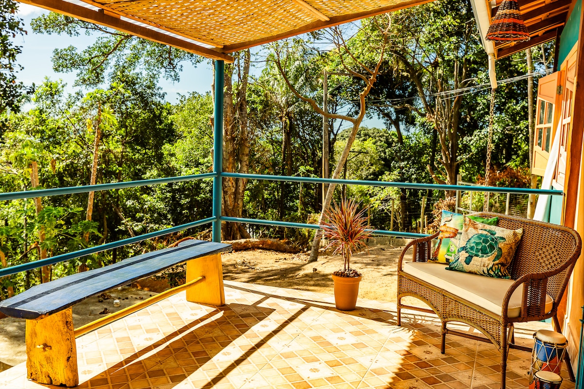 The covered balcony features a wooden bench and a wicker sofa adorned with decorative cushions. A potted plant adds a touch of greenery. In the background, lush trees provide privacy, while glimpses of the sea can be seen through the foliage.