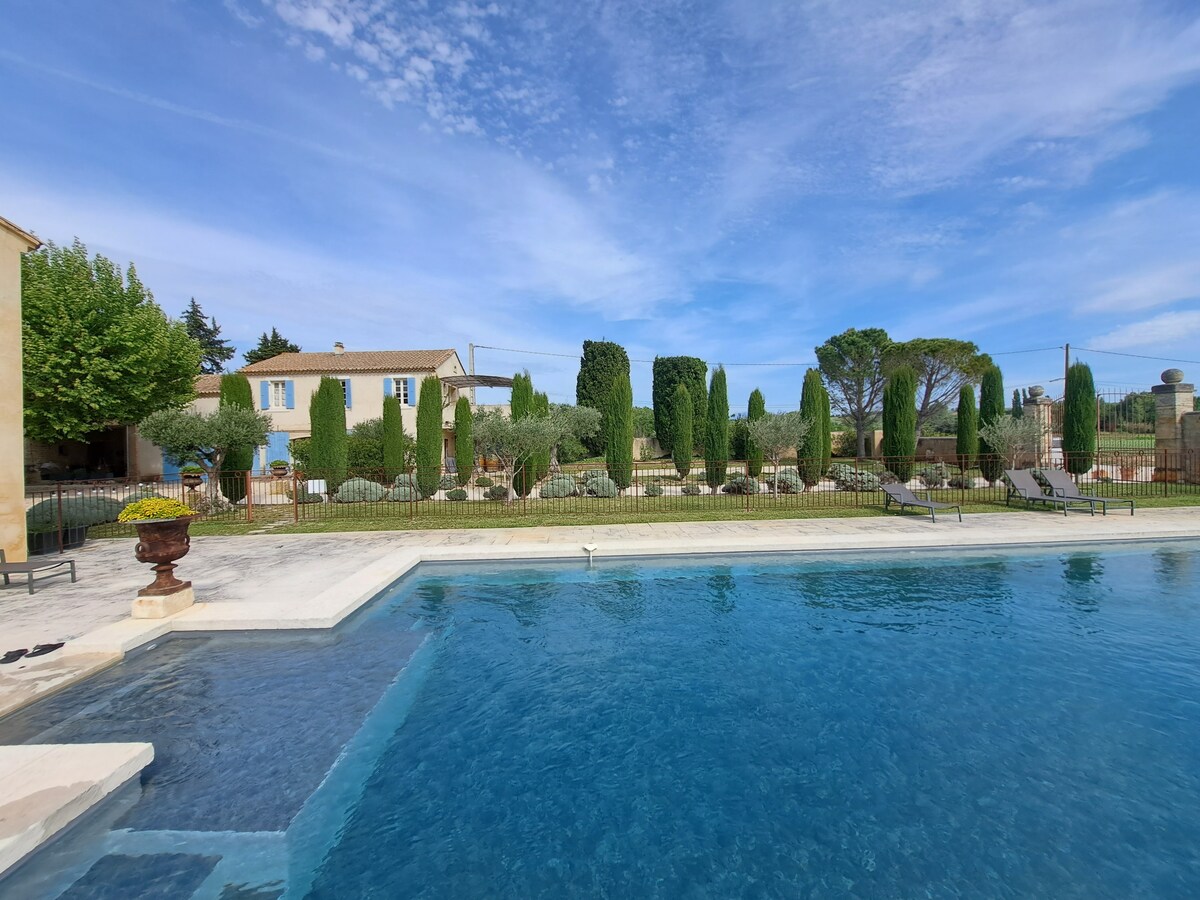 A large, inviting swimming pool is surrounded by a secure fence, with several lounge chairs positioned on the deck. Lush green trees and shrubs are visible in the background, adding to the picturesque landscape of the courtyard.