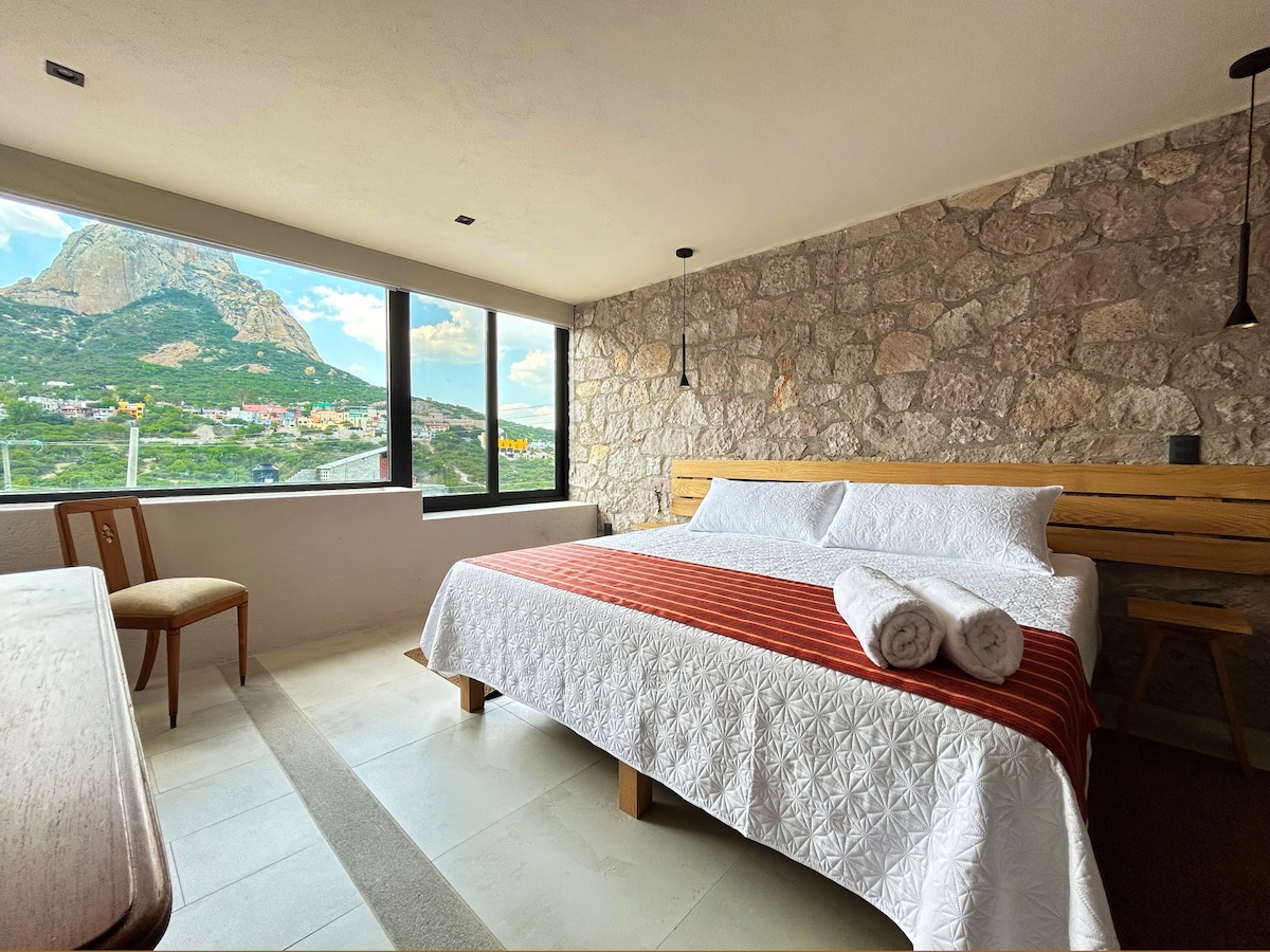 A spacious bedroom features a large bed dressed with white linens and accented by a striped throw. Two towels are neatly arranged at the foot of the bed. Large windows allow natural light to fill the room, showcasing views of Peña de Bernal and the surrounding landscape.
