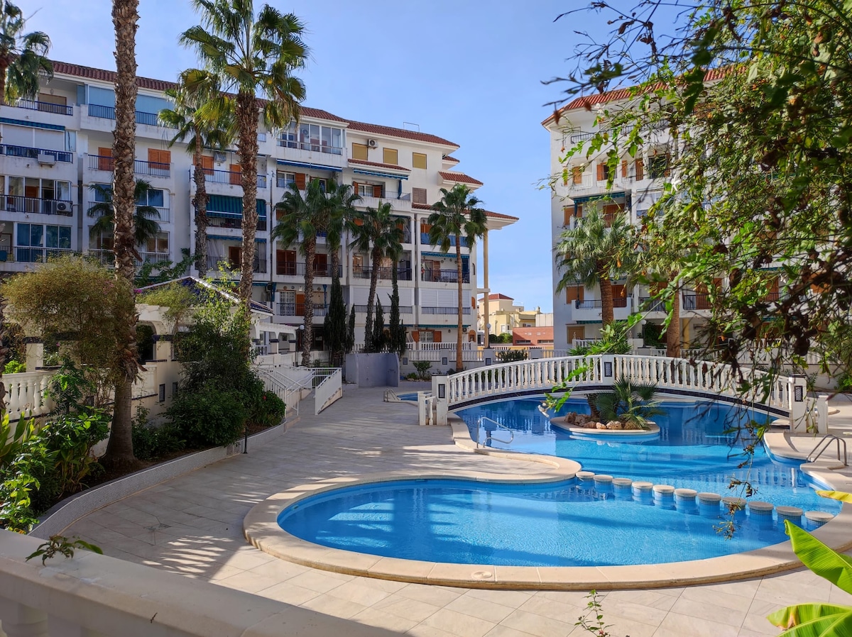 The image depicts a serene pool area surrounded by palm trees and landscaped vegetation. A spacious swimming pool is visible, along with sun loungers and a decorative bridge. The exterior of neighboring buildings reflects a consistent architectural style, contributing to an inviting atmosphere.