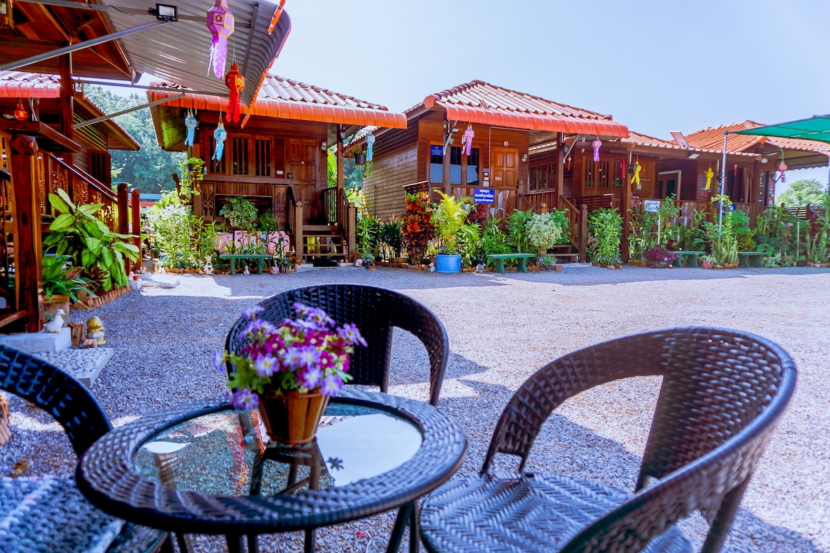 A quaint outdoor seating area features two wicker chairs and a round glass table adorned with a small flower pot. In the background, traditional wooden Thai-style houses are surrounded by lush greenery and decorative plants, contributing to a serene atmosphere.