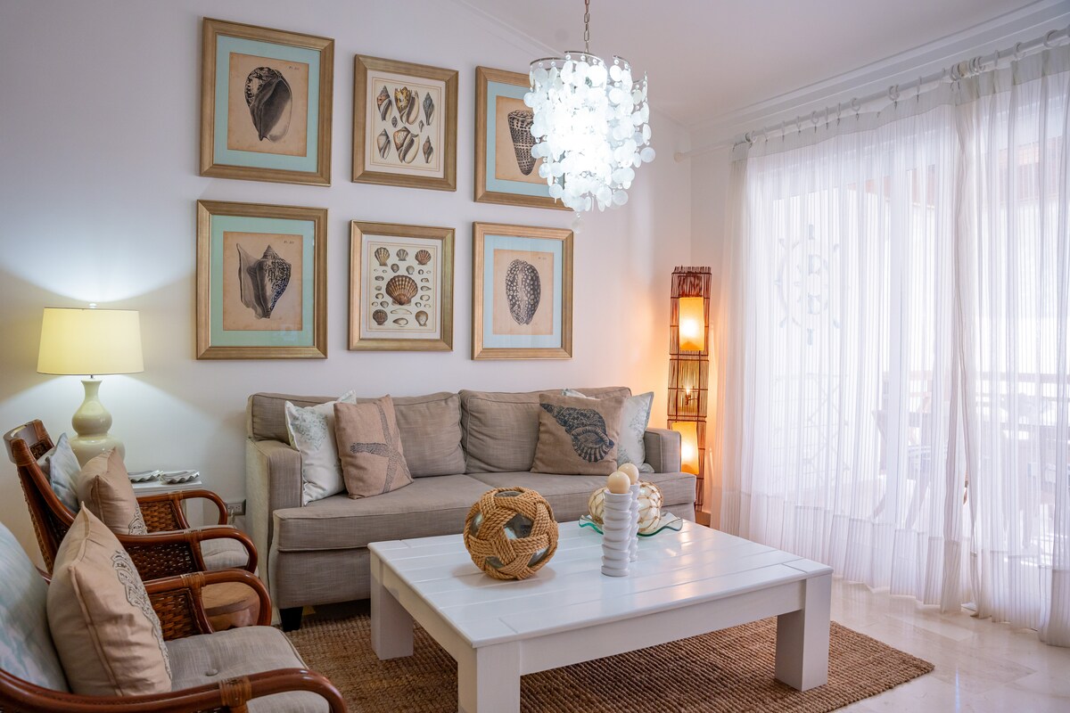 A cozy living room features a light-colored sofa with decorative cushions, complemented by two woven chairs. Unique framed artworks adorn the walls. Soft lighting is provided by a chandelier and lamps, creating a warm atmosphere. A coffee table displays decorative items and fresh flowers.