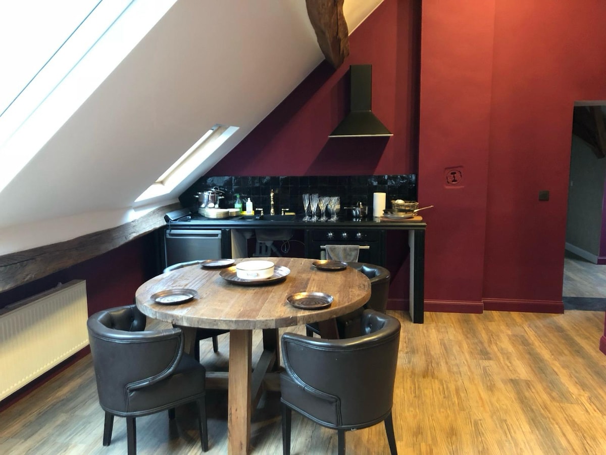 The kitchen area features a round wooden table surrounded by four black chairs. Dark cabinets and a modern stove are visible against a burgundy wall, while overhead windows provide natural light. Cookware and dining items are neatly arranged on the counter.