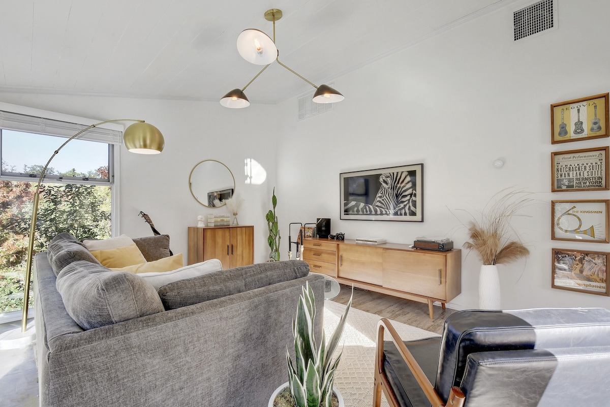 A bright and airy living area showcases a gray sectional sofa positioned near a wooden media console. A round mirror reflects natural light, while decorative plants add a touch of greenery. Artwork hangs on the walls, complementing the space with subtle design elements.