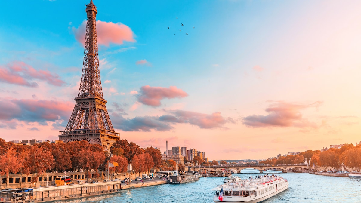 The Eiffel Tower is elegantly framed against a vibrant sky, with hues of pink and orange blending at sunset. The Seine River is visible in the foreground, featuring a boat cruising along the water, while trees and buildings line the riverbank.