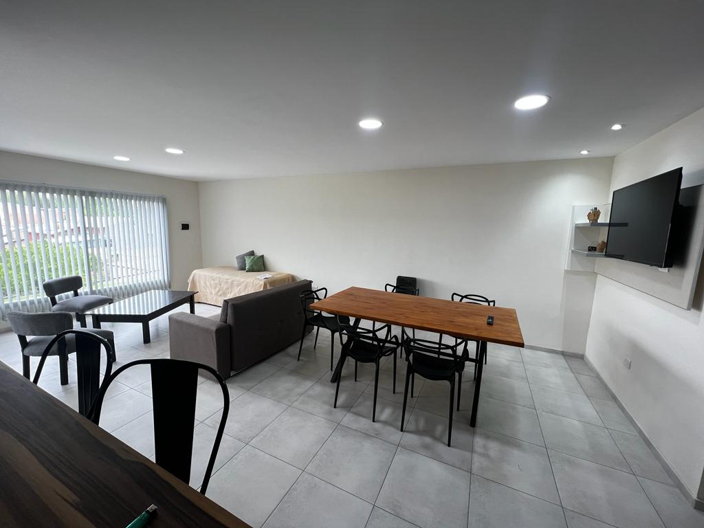 A spacious living area features a comfortable sofa and a large dining table set for several guests. Natural light enters through vertical blinds, illuminating the neutral walls and tiled flooring. A television is mounted on the wall, providing entertainment options.