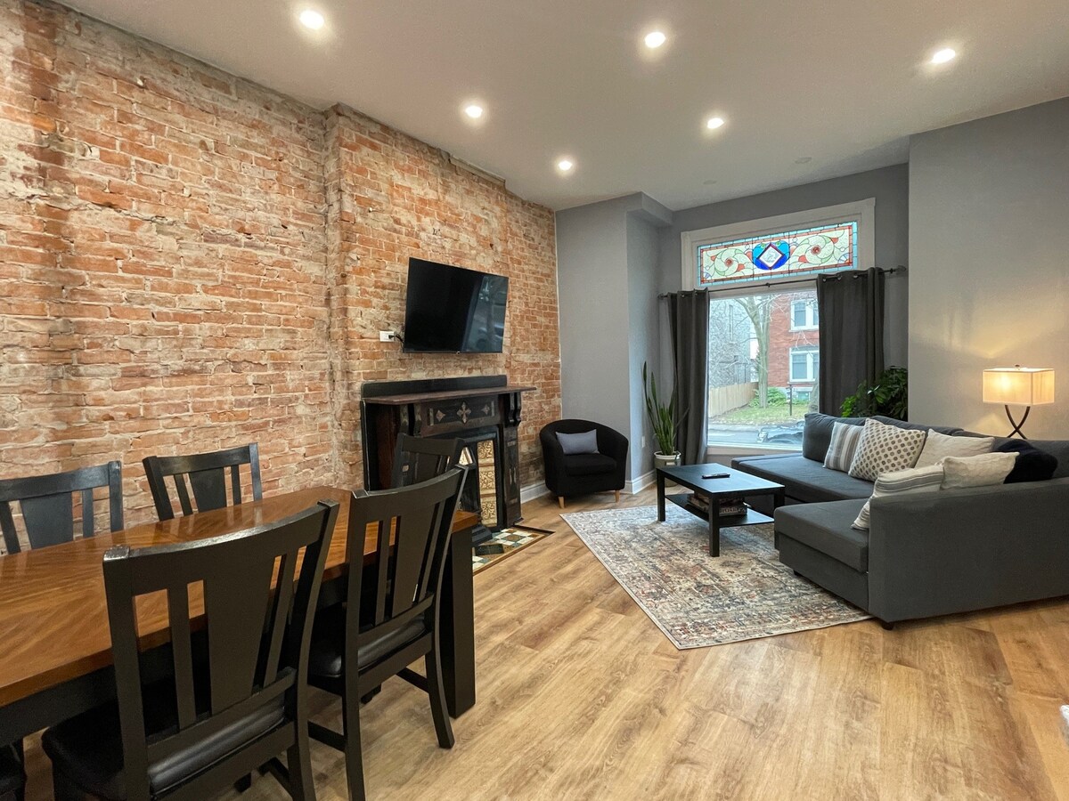 An open living area showcases original exposed brick walls juxtaposed with modern furnishings. A dark wood dining table is positioned beside a cozy seating arrangement, featuring a sectional sofa and a coffee table. Light is provided by recessed ceiling lights and a floor lamp near a window adorned with stained glass.